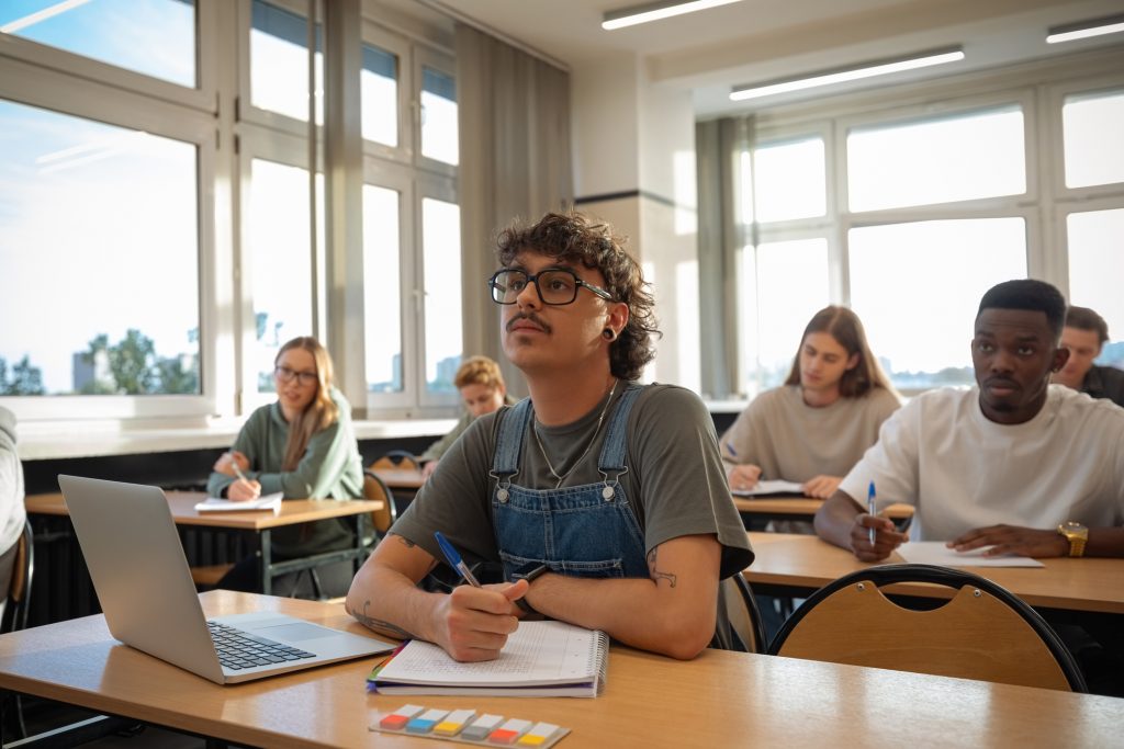 Multiracial group of gen z students sitting in the classroom. Focus on young man using laptop and taking notes.
