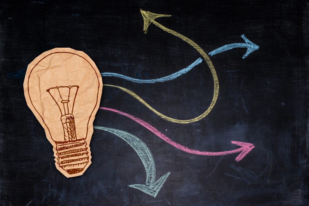 Brown crumpled paper cut in form of a bulb on top of hand drawn multicoloured arrows on blackboard