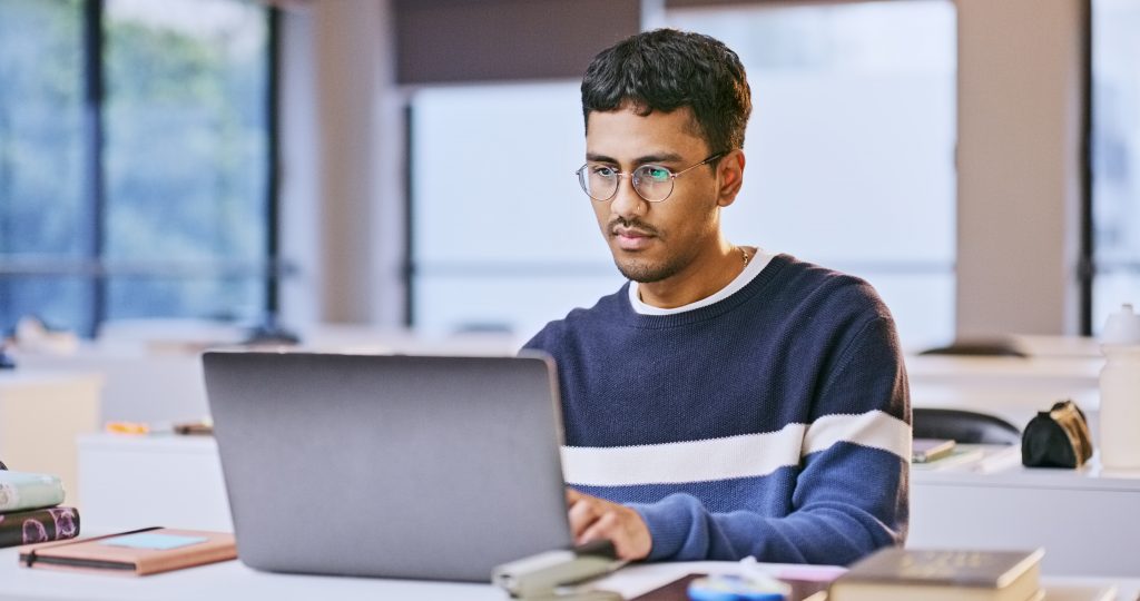 A young Indian male looks thoughtfully at his laptop in a lecture room.