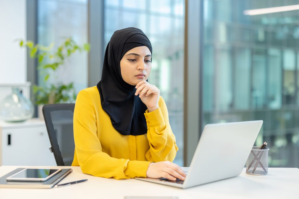 A woman wearing a hijab thoughtfully works on a laptop at her office desk, with a focused expression.